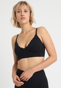 Soutien-gorge noir avec des bords festonnés, en tissu doux, avec des bretelles ajustables et un design sans couture. L'étiquette sur le côté indique "SKNB."