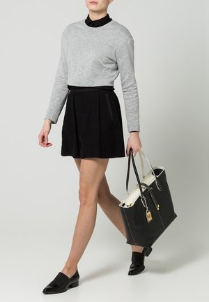 Femme portant un pull gris, une jupe noire et des chaussures noires, tenant un grand sac fourre-tout noir et blanc avec des accents dorés.