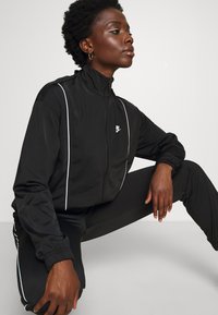 Giacca in felpa nera con zip, dettagli in bianco e un piccolo logo. Presenta un colletto alto e polsini elasticizzati, abbinata a pantaloni neri coordinati.