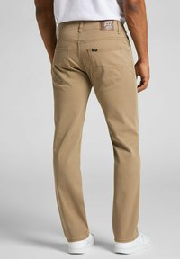 Pantaloni in cotone beige con taglio dritto, dotati di due tasche posteriori e un'etichetta con logo marrone in vita. Dettaglio in tessuto testurizzato.
