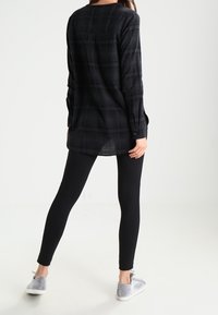 Camicia oversize a quadri neri e grigi con polsini abbottonati, abbinata a leggings neri e scarpe slip-on grigie. Texture del tessuto liscia.