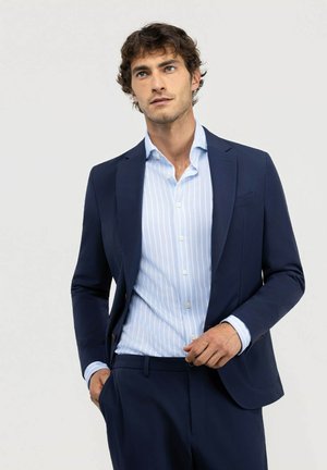 Hombre con traje azul marino y camisa azul claro a rayas, de pie con una mano en el bolsillo, mirando ligeramente hacia arriba y hacia un lado.