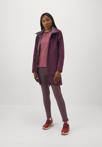 Paarse softshelljack met een fleece-gevoerde kraag, gecombineerd met een roze top en paarse leggings. Rode sneakers maken de outfit compleet.