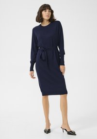 Robe en tricot marine avec des manches longues, une ceinture à la taille et des poignets côtelés. Le tissu est doux et lisse, complété par des talons aiguilles noirs.