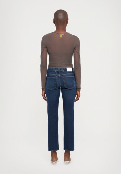 Long-sleeve sheer top in taupe, paired with dark blue straight-leg jeans. Notable details include minimal stitching and a tag at the neckline.