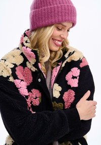Fleecová bunda s kvetinovým vzorom v ružovej, krémovej a zelenej farbe, s čiernym základom, vysokým golierom a pružnými manžetami. V súprave s ružovou pletenou čiapkou.