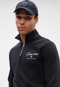 Muž v čiernej mikine s krátkym zipsom a čiernej čiapke s textom "Gant 1949" a logom American Sportswear na hrudi a čiapke.