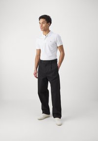 Valkoinen polo-paita, jossa on kaulus, mustat laskostetut housut ja valkoiset lenkkarit. Asusteessa on minimalistinen muotoilu ja sovitettu siluetti.