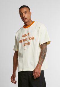 T-shirt en coton crème à manches courtes, col côtelé orange, avec un texte orange audacieux disant "AIN'T HERE FOR LOVE". Coupe standard.