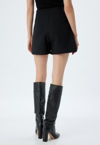 Zwarte op maat gemaakte shorts met een gestructureerde pasvorm, voorzien van twee achterzakken. Gecombineerd met kniehoge zwarte leren laarzen en een aansluitend shirt.