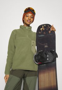 Olivgrüner Fleece-Pullover mit hohem Kragen, schwarzes Snowboard mit gemustertem Oberteil und schwarzen Bindungen. Das Model trägt eine orange Mütze und eine Skibrille.