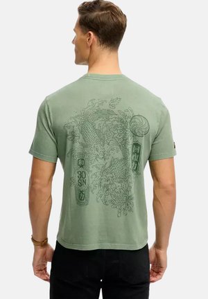 Mand vendt væk iført en grøn t-shirt med detaljeret koi fisk og blomsterdesign trykt på ryggen, kombineret med sorte bukser.