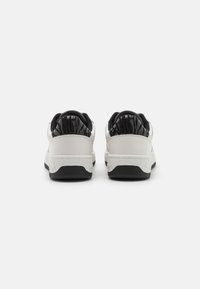 Zapatillas blancas con una parte superior de cuero liso, talón y lengüeta de patrón negro, y suela de goma negra texturizada. Diseño minimalista.