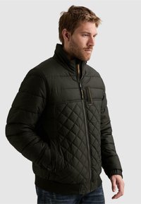 Homme aux cheveux courts et à la barbe, portant une veste matelassée noire zippée avec les mains dans les poches, regardant vers la droite sur un fond clair uni.