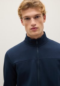 Chaqueta navy de cremallera con collar alto, textura suave y detalle de costura horizontal en el pecho. Diseño minimalista sobre un fondo neutro.