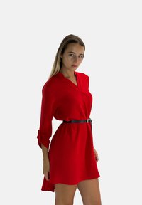Robe rouge avec manches longues, décolleté en V, tissu texturé, cintrée à la taille par une ceinture noire, ourlet fluide et détail des manches retroussées.