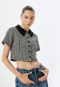Camisa recortada en blanco y negro con estampado de houndstooth, manga corta, gran cuello de cuentas negras y cierre frontal con botones. Combinada con jeans de cintura alta.