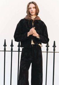 Giacca in corduroy nero con colletto in pile marrone, abbinata a pantaloni in corduroy coordinati. Tessuto testurizzato con una leggera lucentezza.