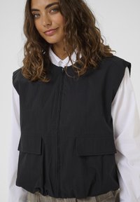 Egy göndör barna hajú nő fekete ujjatlan mellényben áll egy fehér ing és kockás nadrág felett, egy egyszínű háttér előtt.