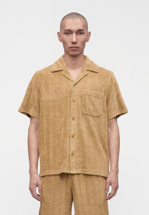 Jeune homme à la tête rasée portant une chemise à manches courtes couleur sable avec boutons et un short assorti sur un fond clair uni.