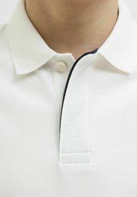 Polo blanc avec un col texturé, une fermeture à un bouton, et une bordure contrastante foncée le long de la patte de boutonnage.