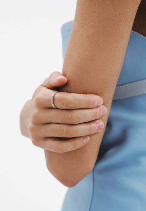 Anillo de plata con un patrón floral en una mano que sostiene un brazo marrón, mostrando la textura natural de la piel contra una prenda azul.