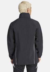 Schwarze wasserdichte Jacke mit hohem Kragen, elastischen Bündchen und weißen seitlichen Streifen. Glatte Textur, strukturierte Passform und Raglanärmel.