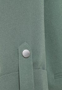 Tissu vert à surface texturée, avec un bouton argenté et un rabat pointu. Idéal pour identifier les caractéristiques du matériau et du design.