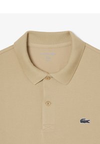 Polo beige con cuello acanalado, tapeta de dos botones y tejido texturizado. Presenta un pequeño logo de cocodrilo negro en el pecho.