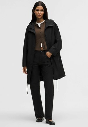 Woman wearing a black hooded coat over a brown buttoned cardigan, black pants, and black flats, standing against a plain light background.