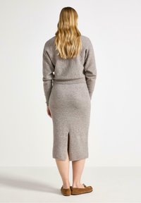 En person med långt, vågigt hår bär en mysig grå tröja och en välsittande midikjol med en slits bak, i kombination med bruna loafers.