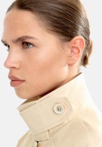 Beżowa kurtka z wysokim kołnierzem o teksturowanym wykończeniu, z widocznym dużym guzikiem i czystym, strukturalnym krojem. Widoczny detal kołnierza.