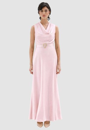 Donna che indossa un abito senza maniche in raso rosa pallido con scollo drappeggiato e vita con cintura, in piedi davanti a uno sfondo chiaro e neutro.