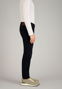 Dunkelblaue Skinny-Jeans mit glatter Textur und Gesäßtaschen, kombiniert mit einem hellen Langarmshirt und grauen Sportschuhen.