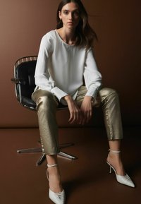 Valkoinen pitkähiha blouse pyöreällä pääntiellä, yhdistettynä metallinhohtoisiin kultaisiin tiukkoihin housuihin ja valkoisiin kärjellisiin korkokenkiin, istuu mustalla tuolilla.