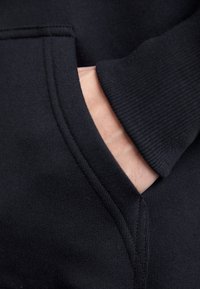 Main partiellement glissée dans la poche avant d’un sweat noir, montrant les détails du tissu et des coutures.