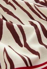 Tissu crème avec de larges rayures irrégulières brun-rouge. Texture douce avec une bordure rouge le long d'un côté.