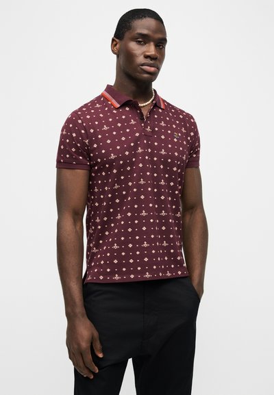 Άνδρας με πουκάμισο πόλο μπορντό με μοτίβο, με κόκκινη και πορτοκαλί περιμετρική λωρίδα στο γιακά και μαύρο παντελόνι, στέκεται μπροστά σε απλό λευκό φόντο.