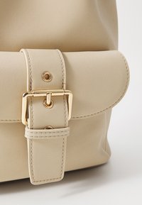 Sac en simili cuir beige avec un rabat enroulé, une boucle carrée dorée et des détails de couture décoratifs. Texture lisse avec un matériel minimal.