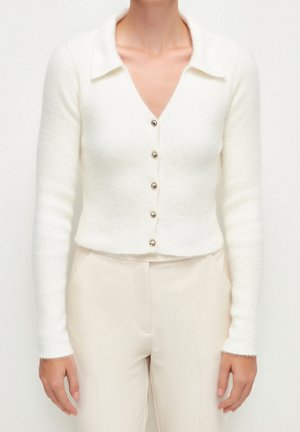 Donna che indossa un cardigan bianco aderente e morbido con bottoni simili a perle e pantaloni beige chiaro abbinati, in piedi davanti a uno sfondo semplice.