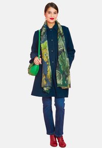 Navy coat with button closure, green floral scarf, blue jeans, and red ankle boots. Green crossbody bag adds color contrast.