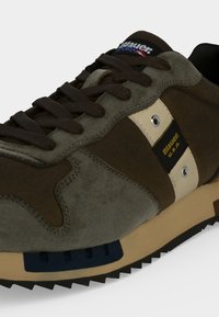 Gros plan sur une basket marron et olive avec des lacets foncés, une bande beige, une étiquette bleue indiquant Blauer U.S.A. et une semelle nervurée.