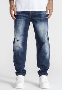 Hombre con jeans azules desgastados y desteñidos y zapatillas blancas, de pie con las manos relajadas a los lados contra un fondo liso.