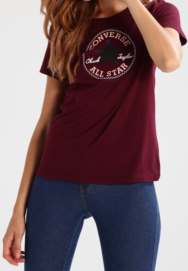 Femme portant un t-shirt Converse All Star bordeaux et un jean bleu, posant avec un bras partiellement levé et les cheveux lâchés.