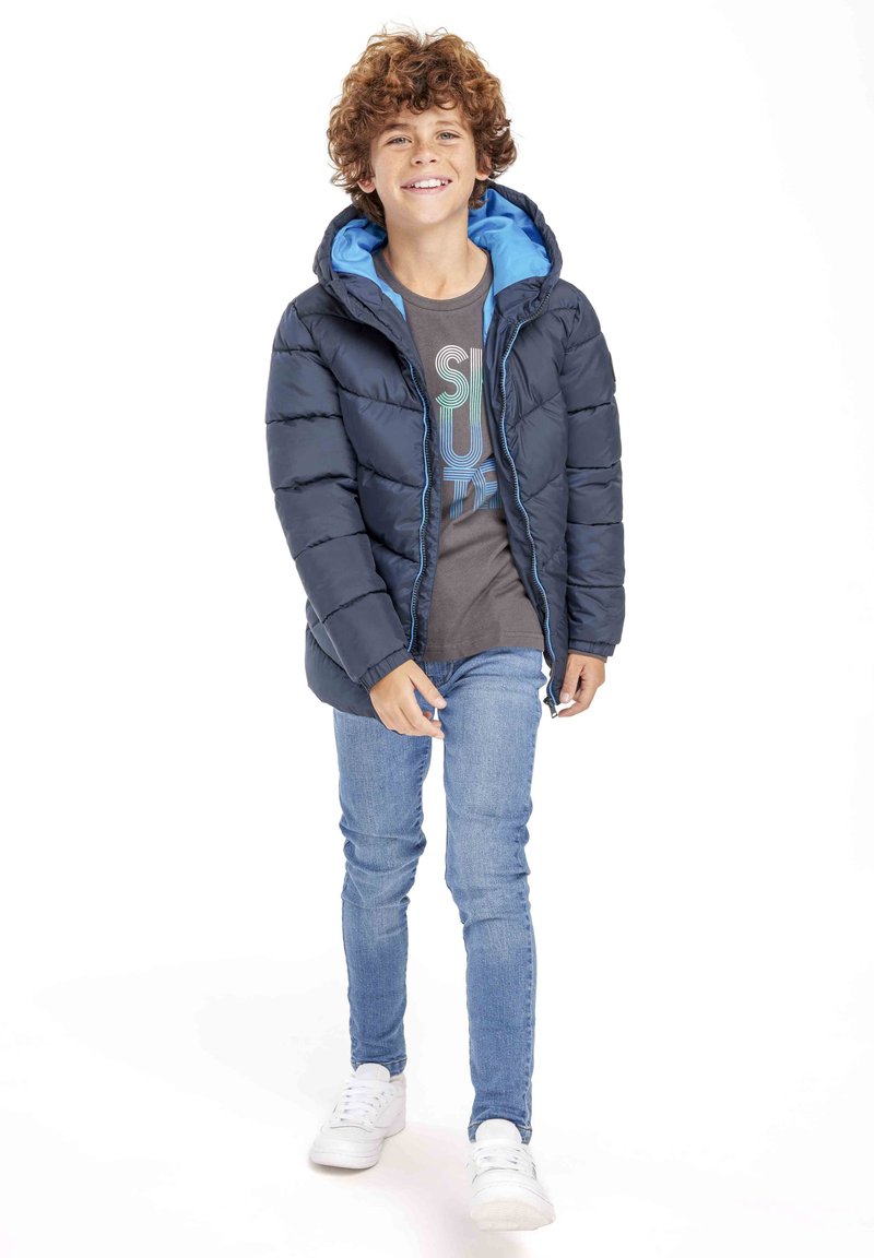 Marineblauwe pufferjack met een blauwe capuchon, voorrits en geweven textuur. Gepaard met een grijs t-shirt en lichtblauwe jeans, en witte schoenen.