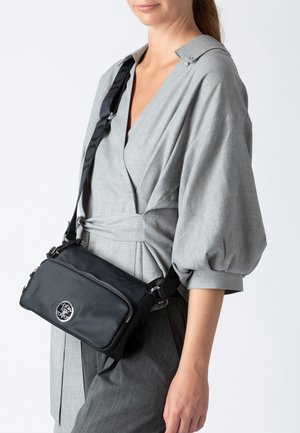 Schwarze Nylon-Umhängetasche mit einer Reißverschlusstasche vorne, kreisförmigem Logodetail und verstellbarem Riemen, getragen über einem grauen Kleidungsstück.