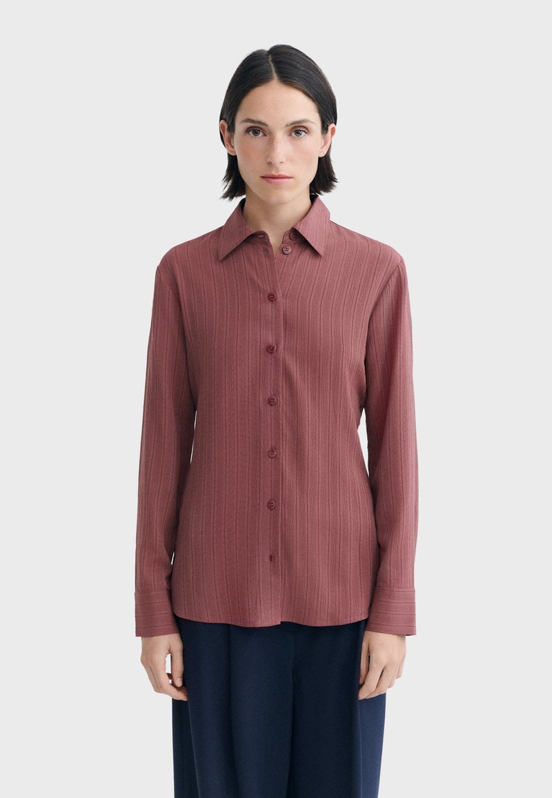 Femme aux cheveux courts et foncés porte une chemise mauve côtelée à manches longues boutonnée et un pantalon foncé, debout devant un fond clair uni.