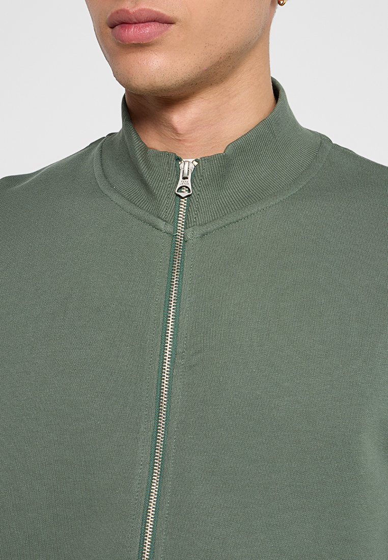 Zielona kurtka zapinana na zamek z wysokim kołnierzem, o prążkowanej teksturze, z metalowym detalem zamka z przodu, wykonana z miękkiego, elastycznego materiału.