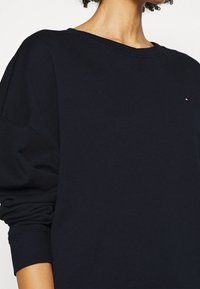 Sudadera de color azul marino con cuello redondo, hombros caídos y puños acanalados. Presenta un pequeño logo decorativo en el pecho. Tejido suave y texturizado.
