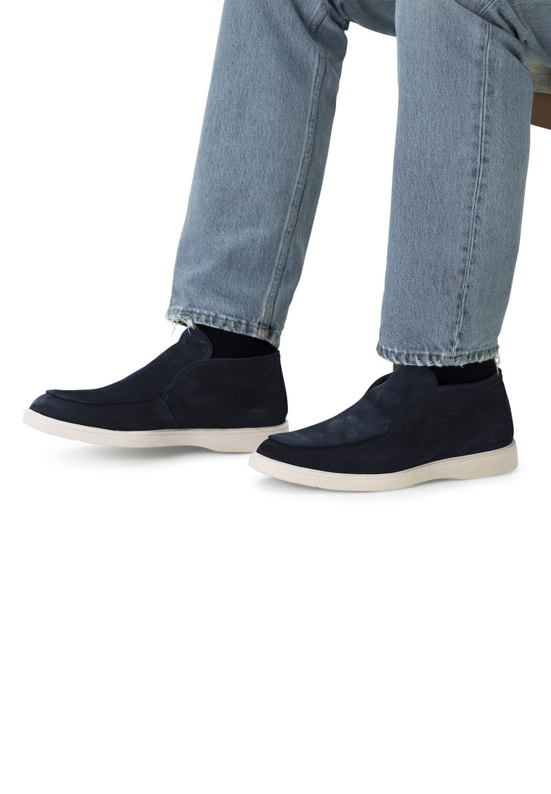 Personne portant un jean bleu clair et des chaussures slip-on bleu foncé avec des semelles blanches, assise les pieds à plat sur le sol.